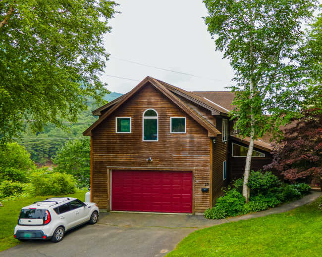 A two-story wooden house with a red garage door is surrounded by trees and situated on a sloped driveway.