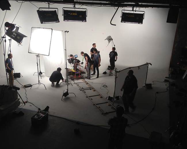 A film crew sets up camera equipment on a studio set with a white backdrop and softbox lights.