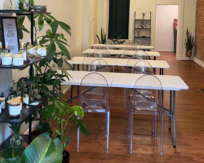 The image shows a spacious room with several white tables arranged for an event, alongside a wall shelf displaying various bottles and plants.