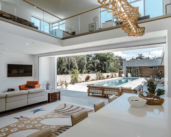 The image shows a modern open-concept living area leading to an outdoor pool, with large sliding glass doors and minimalistic furnishings.