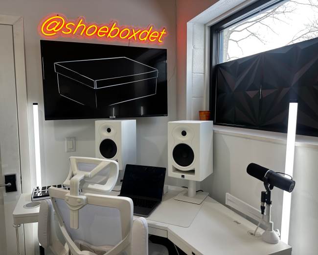 The image shows a modern workspace featuring a desk with a computer, a microphone, and audio speakers, alongside a wall-mounted screen displaying a graphic of a shoebox and a neon sign that reads "@shoeboxdet."