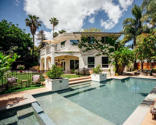 The image shows a two-story house surrounded by green landscaping and a rectangular swimming pool in the foreground.