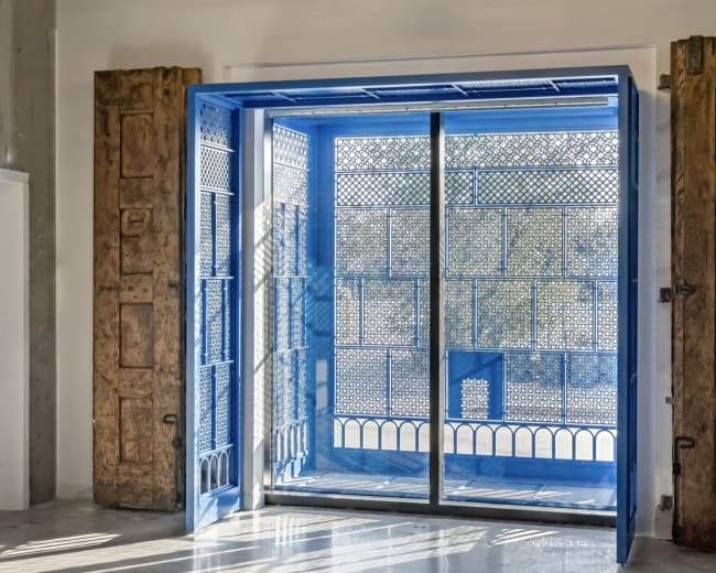 A large blue door with intricate ironwork is open, revealing a bright, sunlit courtyard beyond.