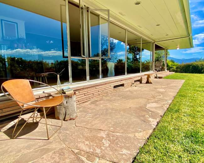 A modern home features a stone patio with a metal chair and large glass windows overlooking a grassy landscape.