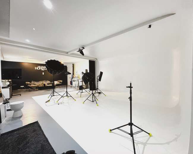 The image shows a photography studio featuring a white backdrop, multiple lighting equipment, and a minimalist setup with a sofa and a dark rug in the corner.
