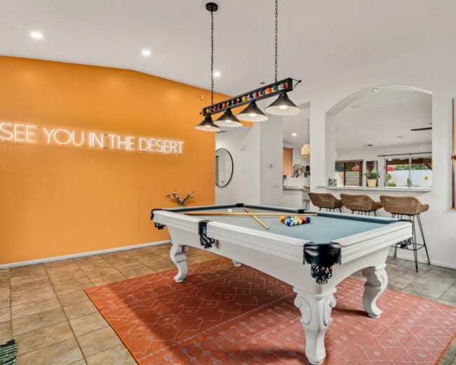 The image shows a game room with a pool table, orange accent wall featuring a neon sign, and bar-style seating in the background.