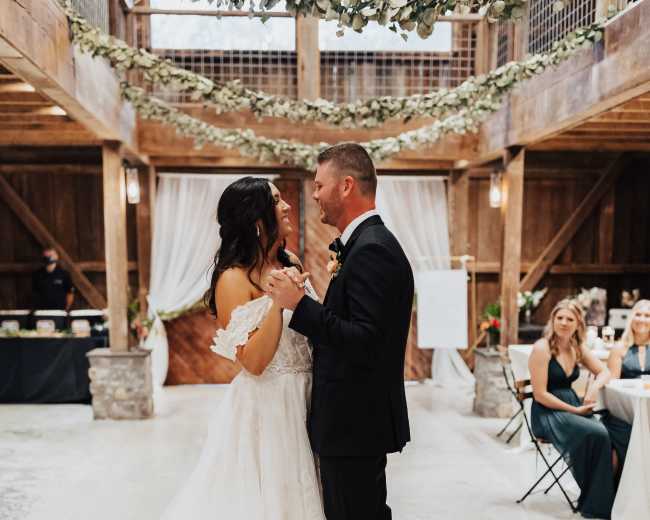 A couple dances together in a rustic barn setting adorned with greenery and floral decorations, while guests watch from seated arrangements.