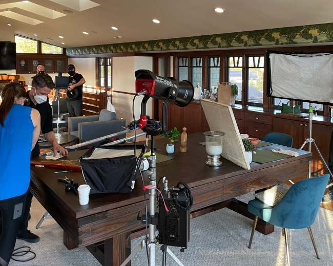 A group of people is preparing a photo shoot in a brightly lit, modern living room with a large wooden table and various equipment set up around it.