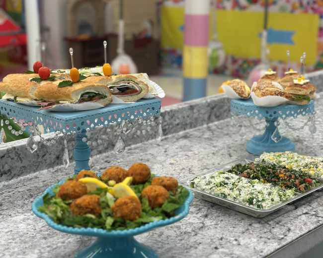 The image shows a colorful buffet setup featuring platters of appetizers, including sandwiches, fried balls, and a salad, arranged on decorative stands.