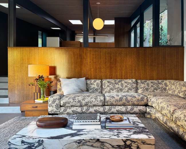 A modern living room features a floral-patterned sectional sofa, a marble coffee table, and warm wooden accents with large windows bringing in natural light.