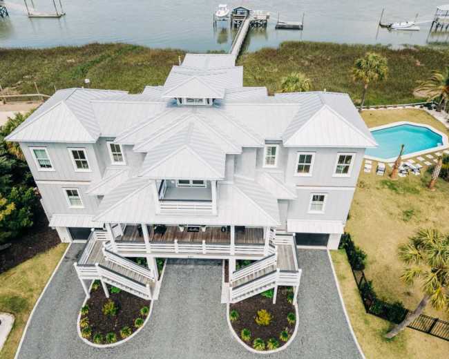 The image shows a large, gray coastal house with multiple levels, surrounded by landscaped grounds, a swimming pool, and a dock extending towards a body of water.