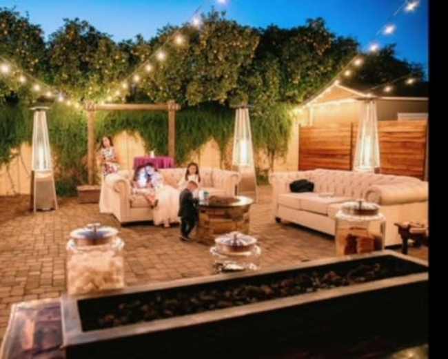 The image shows an outdoor lounge area with string lights, comfortable seating, and decorative plants in the background.