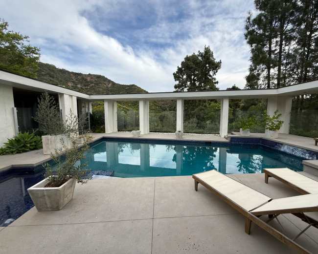 The image shows a modern outdoor swimming pool surrounded by a minimalist patio and lush greenery, with mountains in the background.