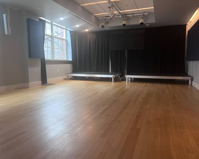 The image shows an empty studio space with wooden flooring, large windows, and dark curtains on the sides.