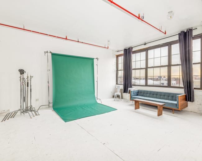 The image shows a spacious, brightly lit studio featuring a green backdrop, a wooden bench, and tall windows.