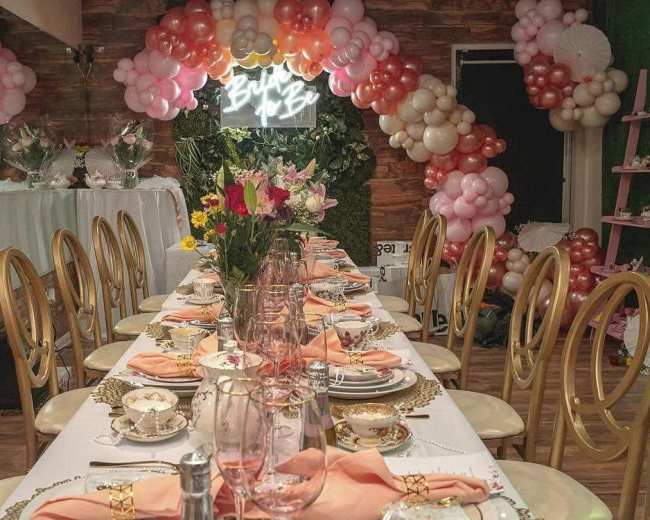 A long dining table is elegantly set with floral centerpieces, place settings, and a backdrop of pink and white balloon decorations.