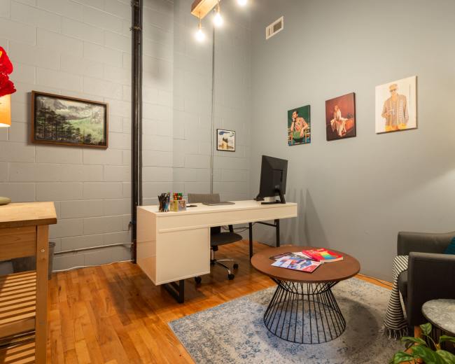 The image shows a small office space featuring a desk with a computer, a round coffee table, a chair, and wall art.