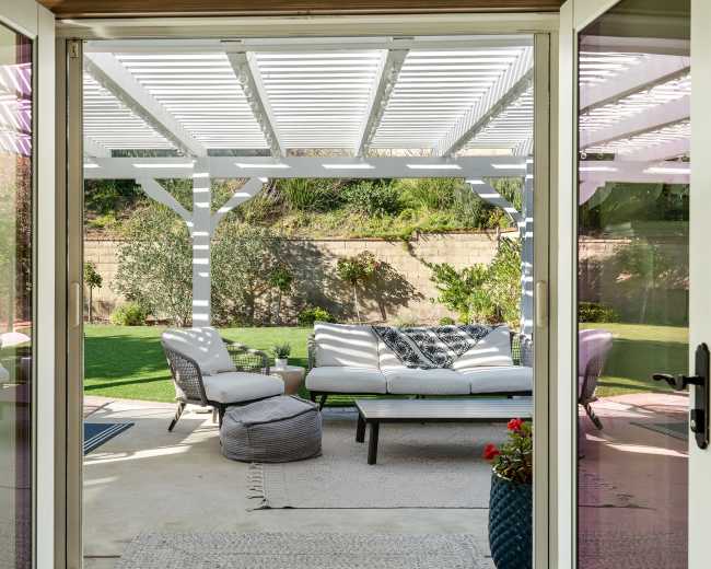 A view from an open doorway reveals a patio with a seating area beneath a white pergola, surrounded by greenery.