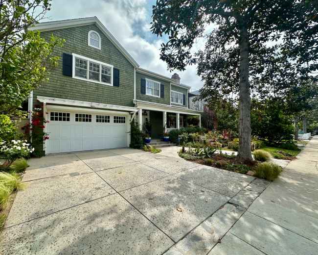 The image shows a large, green-shingled house with a garage and a well-maintained front yard featuring various plants and trees.