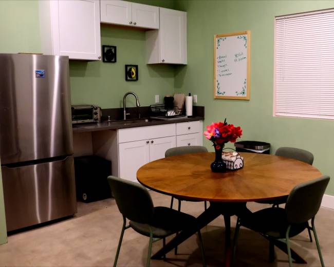 The image shows a kitchen and dining area featuring a round wooden table with four chairs, a metal fridge, and green walls adorned with a whiteboard and a floral centerpiece.