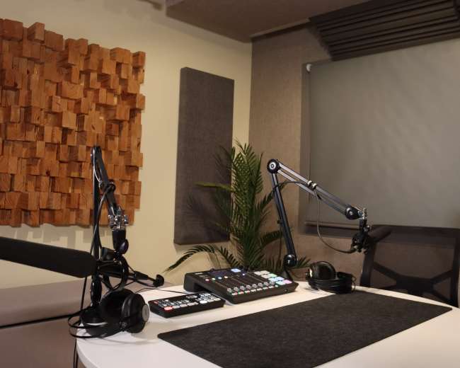 The image shows a modern radio studio with a circular white table, audio equipment, microphones on adjustable arms, headphones, and soundproofed walls.