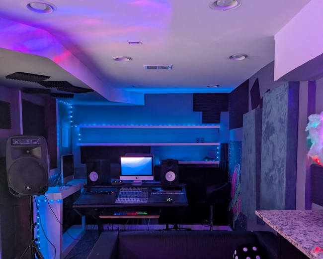 The image shows a modern music studio featuring a mixing desk, speakers, a computer setup, and ambient lighting in shades of blue and purple.