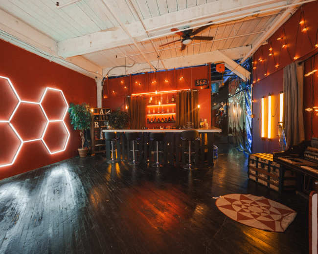The image shows a modern interior space featuring a bar area with hexagonal neon lights on the wall, dim lighting, and a wooden floor.
