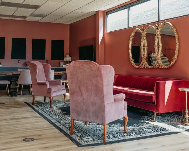 The image shows a modern lounge area with pink chairs, a red sofa, a decorative mirror, and wooden flooring.