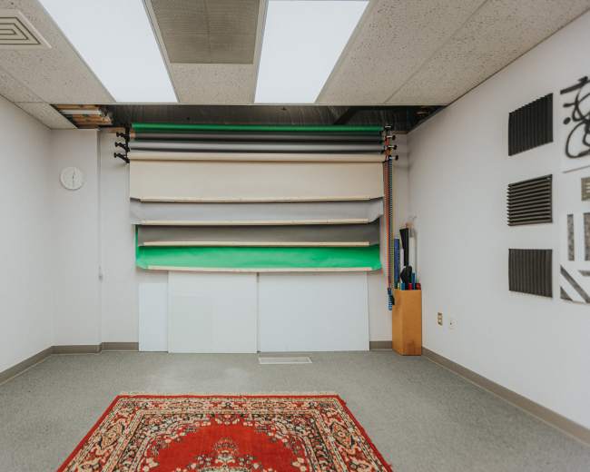 The image depicts a photography studio with a backdrop system of folded rolls in various colors against a white wall, accompanied by soundproofing panels and a decorative rug on the floor.