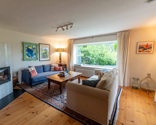 A cozy living room featuring a blue sofa, a beige couch, a wooden coffee table, and a fireplace, with large windows providing a view of greenery outside.