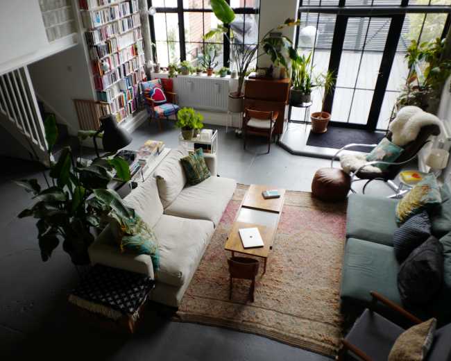 The image shows a spacious living room filled with plants, a large sofa, a coffee table, and a bookshelf, with large windows allowing natural light to enter.