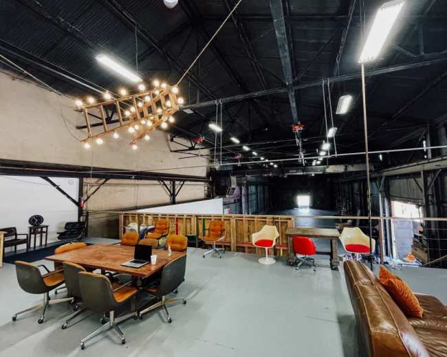 The image shows a spacious, industrial-style loft with a wooden table surrounded by chairs and a sofa, illuminated by a striking chandelier.