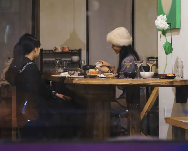 Two people sit at a wooden table in a softly lit room, with one person focused on a dish while the other looks on.