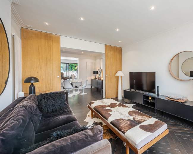 The image shows a modern living room with a dark sofa, a coffee table, a television on a black media unit, and a large round mirror on the wall.