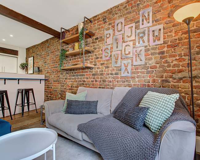 A cozy living room features a gray sofa with patterned cushions, a brick wall, and wooden shelving displaying decorative items.
