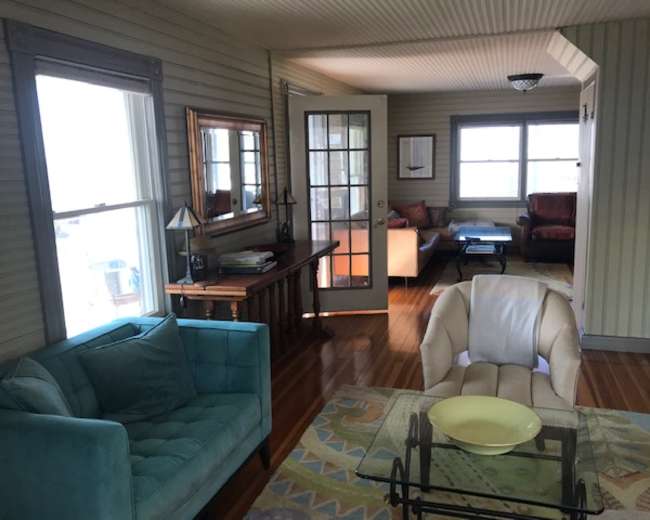 The scene depicts a cozy living area with a teal couch, a cream-colored armchair, a coffee table, and large windows allowing natural light.