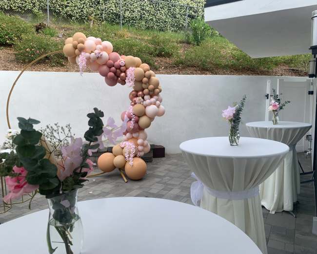 The scene features a decorated outdoor area with round tables draped in white cloth, each topped with a small vase of flowers, and a colorful balloon arch in the background.