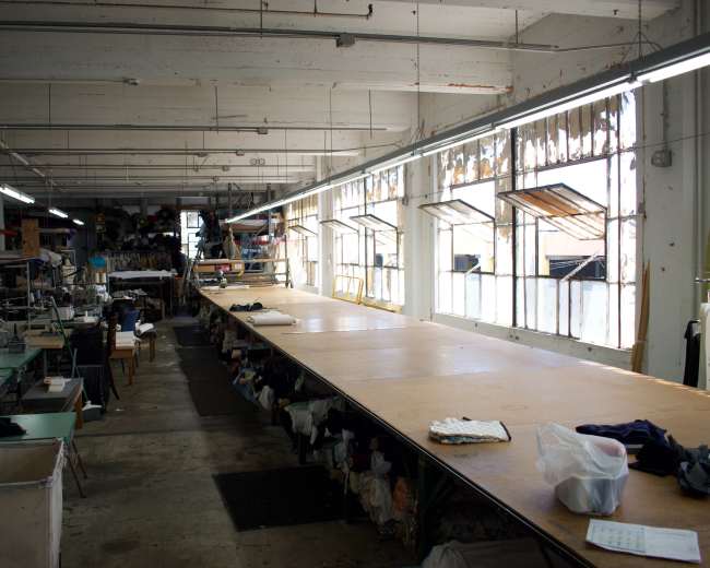 The image shows a large, open workshop space with wooden tables and sewing machines, illuminated by natural light through multiple windows.