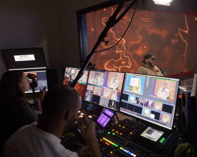 The image shows a busy television studio control room with multiple screens and a crew operating equipment while two hosts are visible on a set in the background.