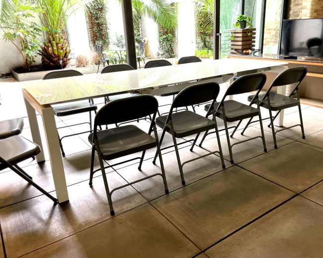 A long, white dining table with several gray folding chairs is positioned in a room with large windows and potted plants.