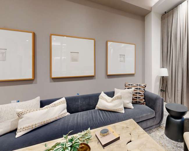 A modern living room features a gray sofa with decorative pillows, a marble coffee table, and framed artworks on the wall, with large windows and light curtains.