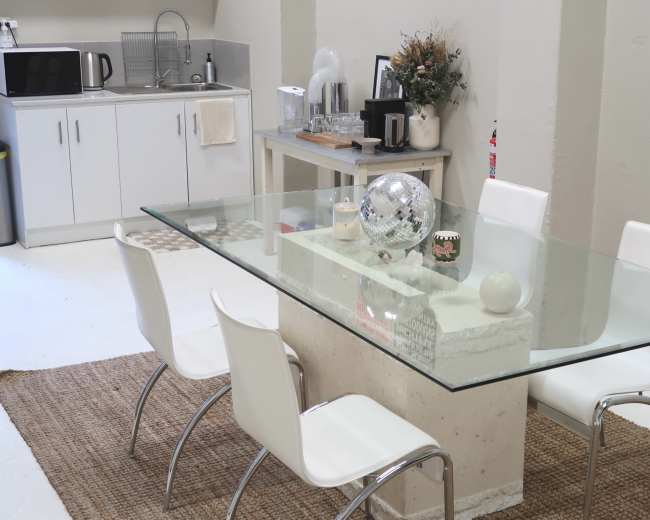 The image shows a dining area with a glass table surrounded by white chairs, a kitchen counter in the background, and various kitchen appliances.