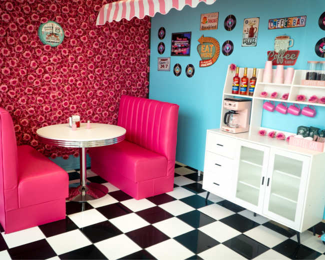 The image depicts a retro-style diner interior with pink booths, a circular white table, and a decorative wall featuring a floral pattern and various retro signs.