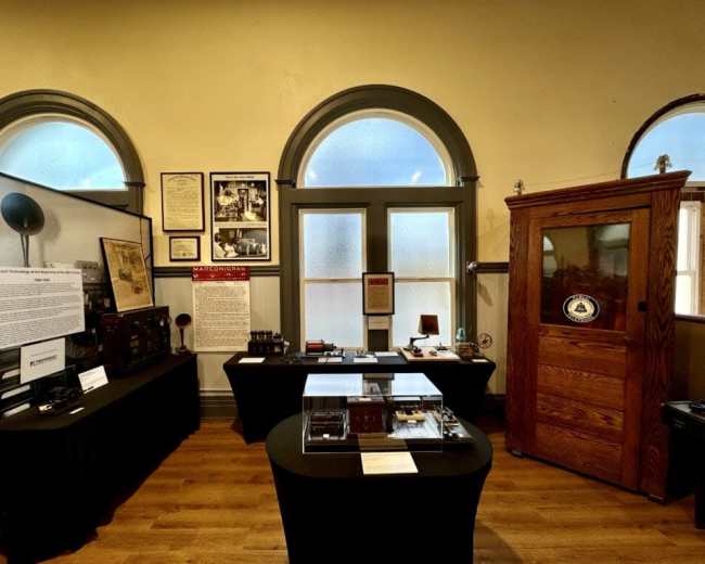 The image shows an exhibit space featuring displays of historical artifacts and informational panels, with large windows providing natural light.