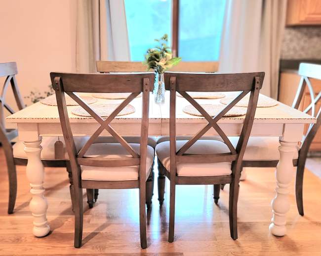 A white dining table set with four wooden chairs sits in a well-lit room near a window, complemented by a small vase of flowers.