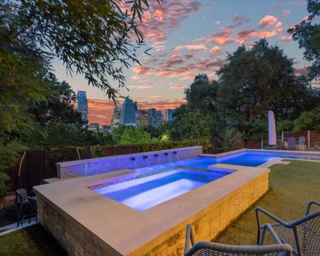 A modern backyard features a rectangular pool with a spa, surrounded by lush greenery and a city skyline at sunset.