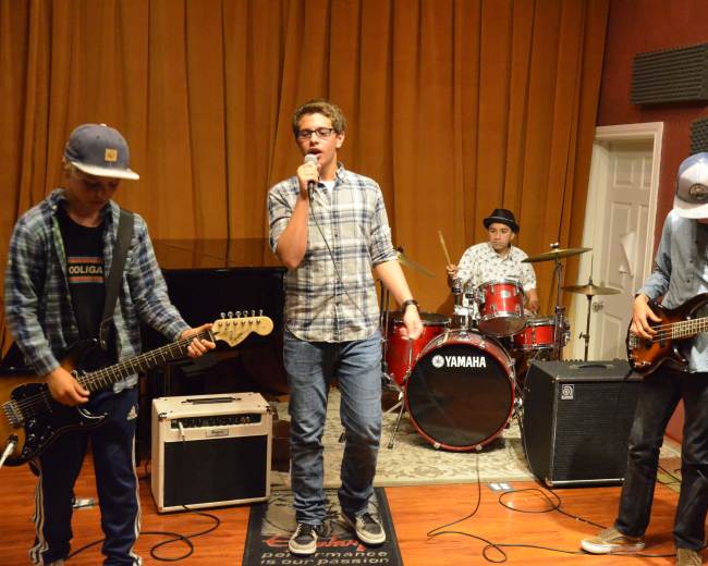 A group of four musicians performs in a studio, with one singer holding a microphone while the others play guitar, bass, and drums.