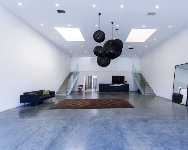 The image shows a spacious, minimalistic interior with polished concrete floors, a large area rug, and a series of hanging black light fixtures above a modern seating area and an open staircase.