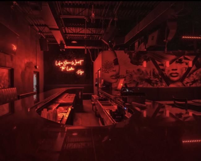 The image shows a dimly-lit bar interior featuring a curved counter, neon signage, and vibrant wall murals.