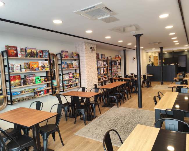 The image shows a game café with rows of tables, shelves filled with board games on the walls, and a counter in the background.